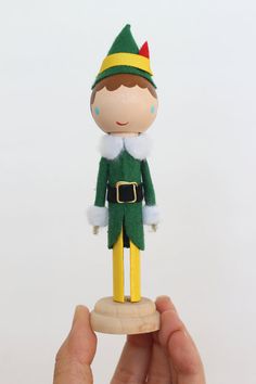 a hand holding a small wooden toy with a green outfit and hat on it's head