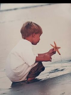 a young boy kneeling down on the beach holding a starfish in his right hand