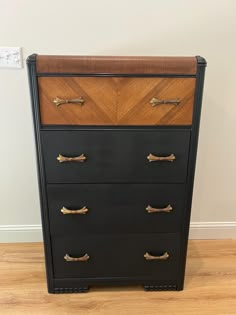 a black and brown chest of drawers with brass handles on the top, in front of a white wall