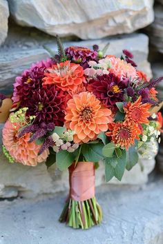 a bridal bouquet with orange and purple flowers in front of a rock wall on a sunny day