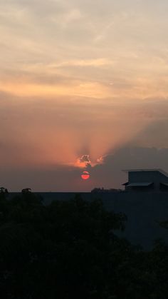 the sun is setting behind some clouds in the sky over a building and tree line