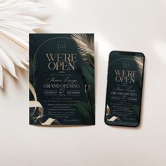 a phone next to a black and gold wedding card on a white table with palm leaves