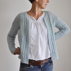 a woman standing in front of a white wall wearing a blue cardigan sweater and jeans