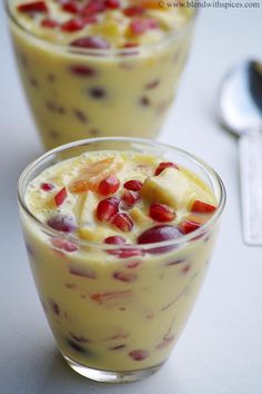 two glasses filled with fruit and pudding on top of a white table next to a spoon