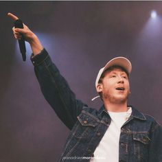 a man in a denim jacket and hat holds his hand up to the sky while standing on stage