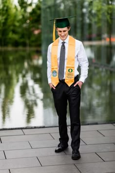 a man in a graduation cap and gown is standing on the sidewalk near water with his hands in his pockets