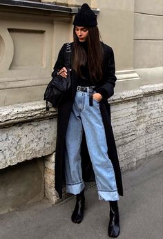 a woman in black coat and jeans leaning against a wall