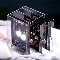 three clear jewelry boxes with earrings in them on a white cloth covered table next to a plant