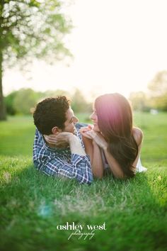 a man and woman laying on the grass in front of trees with their arms around each other