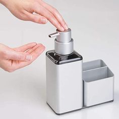 two hands reaching for a soap dispenser on a white surface with one hand holding the soap dispenser