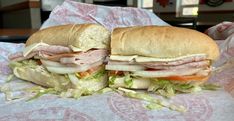 two sub sandwiches cut in half sitting on top of a wrapper with lettuce and tomato