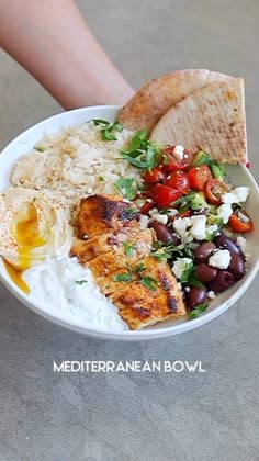 a white bowl filled with rice, beans and other food next to a person's hand