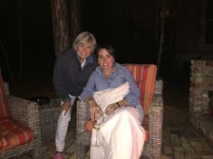 two women sitting on wicker chairs in front of a fire place at night, one holding a wine glass and the other smiling