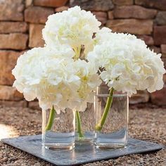 three clear vases with white flowers in them on a stone slab near a brick wall