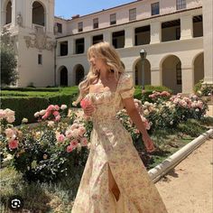 a woman in a long dress is walking through the garden with flowers and bushes behind her