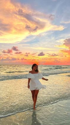 a woman in white dress walking on the beach at sunset with clouds and water behind her