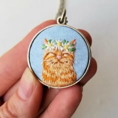 a hand holding a small embroidered pendant with a cat on it's face and flowers in its hair