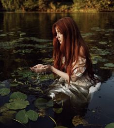 a woman with long red hair sitting in the water surrounded by lily pads and leaves