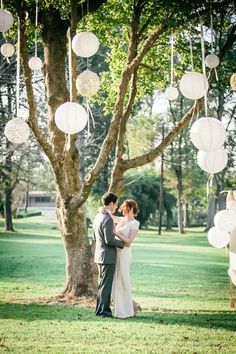 a bride and groom standing under a tree with paper lanterns hanging from it's branches