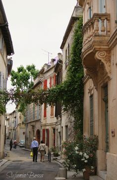 two people walking down the street in front of some buildings with flowers growing on them