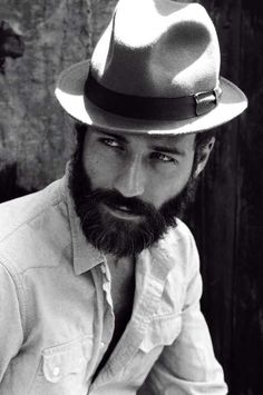 a man with a beard wearing a fedora and white shirt sitting on a bench