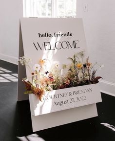 a welcome sign with flowers in it sitting on the floor next to a white wall