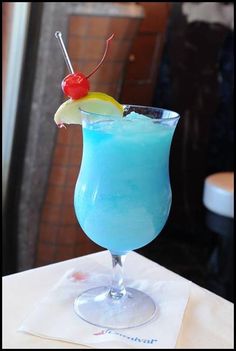 a blue drink sitting on top of a table next to a glass with a cherry in it