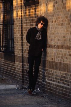 a man leaning against a brick wall with his hands on his hips wearing sunglasses and a scarf