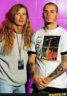 two men standing next to each other in front of a purple wall with tattoos on their arms