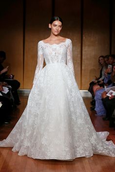 a white wedding dress with long sleeves and sheer lace on the shoulders, is shown in front of a crowd