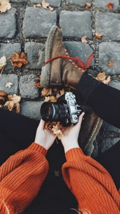 someone holding a camera in their hands while sitting on the ground with leaves around them