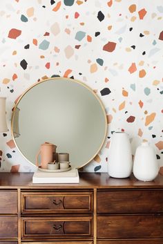 a dresser with a mirror, vases and lamp on it in front of a wallpapered background