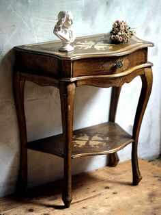 an old wooden table with a vase on top and a small shelf below it, against a white wall