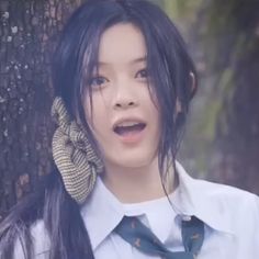 a woman with long black hair wearing a tie and dress shirt, standing in front of a tree