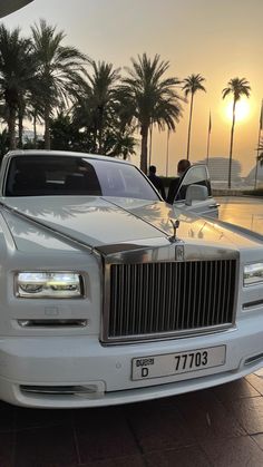 a white rolls royce parked in front of palm trees at sunset with the sun setting behind it