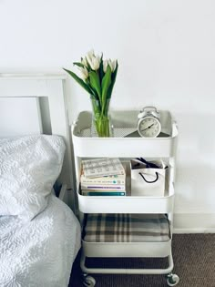 a white shelf with magazines and flowers on it next to a bed in a bedroom