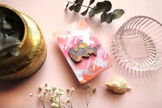 a pink and white card with a cartoon character on it next to some flowers, a glass vase and a gold plate