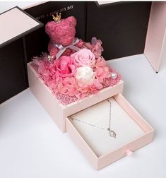 a pink teddy bear sitting on top of a box filled with flowers and a necklace