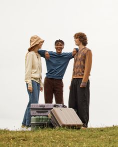 three people standing on top of a grass covered hill next to suitcases and boxes