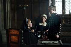 three people dressed in period costumes standing around a table with a woman sitting on it