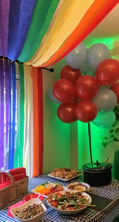 a table topped with plates of food and balloons in the air above it is a rainbow colored wall