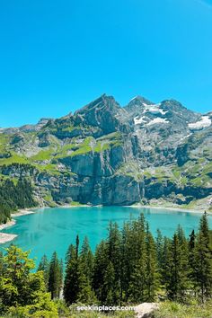 a mountain lake surrounded by pine trees and snow capped mountains in the distance with blue sky