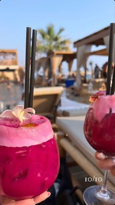 two people holding up drinks in front of the camera with palm trees in the background