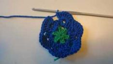 a crocheted blue flower with green leaves sits next to a knitting needle on a white surface