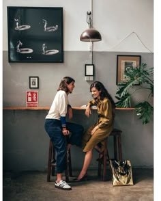 two women sitting at a bar talking to each other in front of a wall with pictures on it