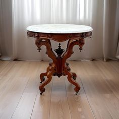 an antique table with marble top in front of a white curtained window and wooden floor