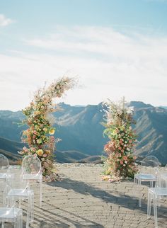 an outdoor ceremony set up with clear chairs and flowers on the arch over looking mountains