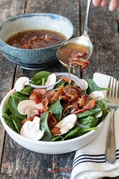 a bowl filled with spinach and bacon on top of a wooden table next to a spoon