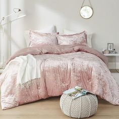 a bed with pink comforter and pillows in a white room next to a round ottoman