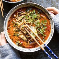 two hands holding chopsticks over a bowl of soup with noodles, mushrooms and vegetables